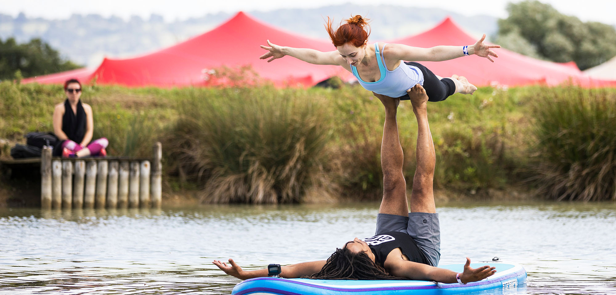 couple going yoga pose on paddle board on lake with red stretch tent in background