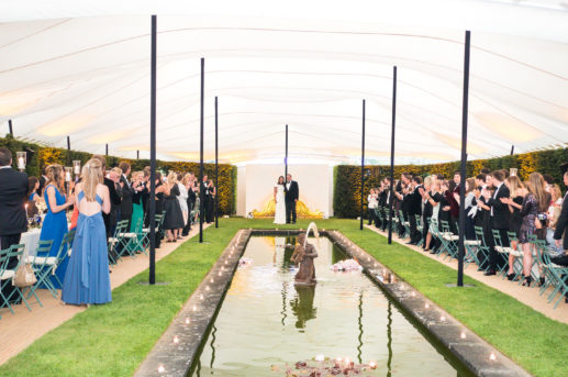 white stretch tent over ornamental pool and hedges with guests on either side standing clapping bride and groom in the middle