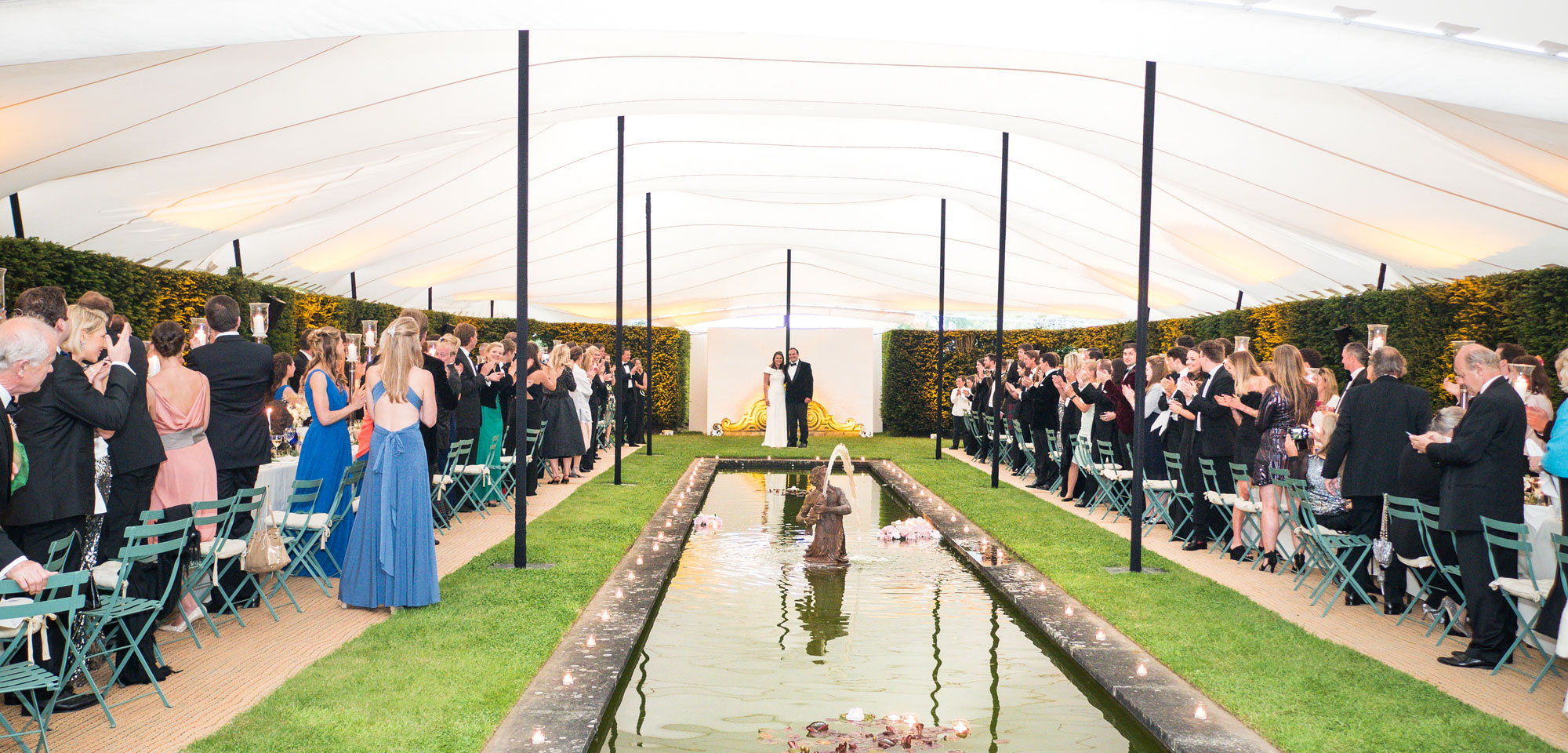 white stretch tent over ornamental pool and hedges with guests on either side standing clapping bride and groom in the middle